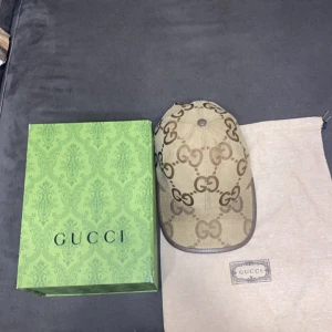 Beige keps från Gucci med GG-mönster - Snygg beige keps från Gucci med klassiskt GG-monogram i brunt. Kepsen har justerbar rem bak i brunt skinn och är tillverkad i canvas. Kommer med originalbox och dustbag. Perfekt för dig som vill ha en lyxig och trendig accessoar.