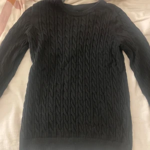 Svart kabelstickad tröja - Svart kabelstickad tröja med rund hals och långa ärmar. Tröjan har ett klassiskt flätmönster över hela plagget och är gjord i ett mjukt stickat material