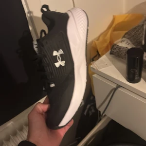 Under Armour Charged Commit TR 4 sneakers - Svarta och vita sneakers från Under Armour, modell Charged Commit TR 4. Skorna har en sportig look med vit sula, snörning och tydlig logga på sidan. Perfekta för träning eller gymmet, med bekväm passform och slitstarkt syntetmaterial.