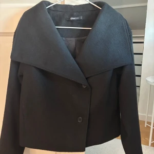 Svart kappa från Gina Tricot XS - Svart kappa från Gina Tricot i kortare modell med bred krage och två knappar framtill. Jackan är tillverkad i ett mjukt ullblandat material. .