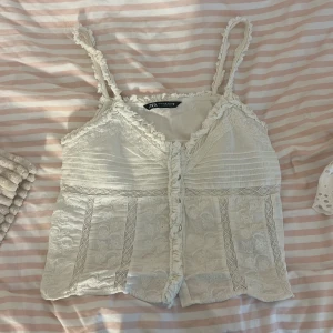 Vit spetsig linnetopp från Zara - Superfin vit linnetopp från Zara med tunna axelband och volangkant. Toppen har broderade spetsdetaljer, dekorativa sömmar och knappar framtill. Perfekt för en somrig look med romantiska vibbar och vintagekänsla. Storlek M men passar mer som S