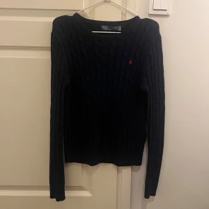Mörkblå kabelstickad tröja Polo Ralph Lauren - Kabelstickad mörkblå tröja från Polo Ralph Lauren i mjuk pima-bomull. Använd en gång. Klassisk rund halsringning, långärmad modell och ikonisk röd broderad logga på bröstet. Perfekt för dig som gillar stilren och tidlös design.