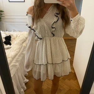 Vit prickig klänning med volang - Supersöt vit kortklänning med svarta prickar, omlott och volangdetaljer. Klänningen har  långa ärmar, samt två lager med volanger i kjolen. Perfekt för dig som gillar romantisk och lekfull stil. Passar perfekt till konfrontation eller student!🤍🤍🩷