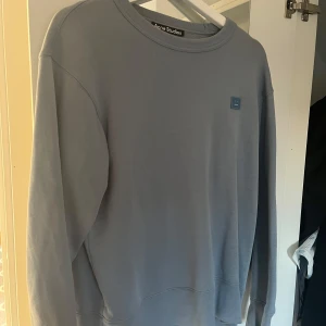 Blå sweatshirt från Acne Studios - Säljer en stilren ljusblå sweatshirt från Acne Studios med deras ikoniska face-logga på bröstet. Tröjan har rund halsringning, långa ärmar och är gjord i mjuk bomull. Väldigt fin!