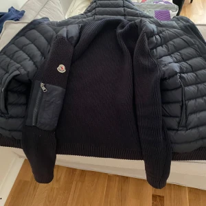 Svart pufferjacka från Moncler - Snygg svart pufferjacka från Moncler med stickade ärmar och detaljerade fickor med dragkedja. Jackan har ribbstickad mudd och klassisk Moncler-logga på ärmen. Perfekt för dig som vill ha en stilren och varm jacka med coola materialmixar.