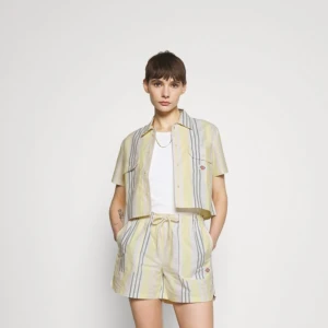 Randig kortärmad skjorta från Dickies - Snygg kortärmad skjorta från Dickies i ljusgult, vitt och grått med vertikala ränder. Modellen är croppad med två bröstfickor och klassisk krage. Skjortan har knappar framtill och logopatch på ena fickan. Perfekt för dig som gillar street och chill stil.