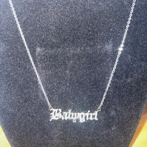 Silvrigt halsband med Babygirl-text - Snyggt halsband i silverfärgad metall med kedja och hängande texten 'Babygirl' i gotisk stil. Perfekt statement-piece för att lyfta din outfit och ge den en cool vibe. Passar dig som gillar trendiga accessoarer med attityd.