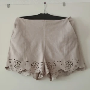 Beige mockashorts med broderad kant - Supersnygga beige shorts i mockaimitation med dragkedja i sidan. Nederkanten har ett unikt broderat mönster med utskurna detaljer och vågig form. 