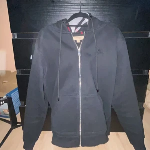 Svart Burberry hoodie med dragkedja - Hej alla säljer en Svart hoodie från Burberry med klassiskt rutigt foder i huvan. Tröjan har dragkedja framtill, snörning vid huvan och två fickor. Tillverkad i mjuk bomull med en clean look och diskret Burberry-logga på bröstet. Perfekt för dig som gillar stilren streetwear. Det är bara och skriva om du har någon fråga.😁