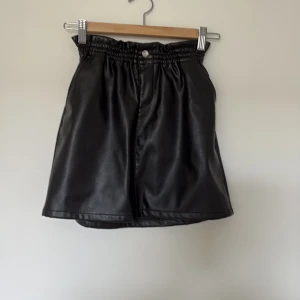 Svart kortkjol i skinnimitation Name It - Cool svart kortkjol från Name It i skinnimitation med elastisk midja och knapp framtill. Kjolen har en lätt A-linjeformad siluett och är perfekt för dig som gillar en edgy stil. Passar till allt från sneakers till boots.