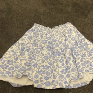 Blommig kortkjol från Zara - Supersöt kortkjol från Zara med blått och vitt blommönster. Kjolen har volanger och resår i midjan för skön passform. Perfekt för dig som gillar ljusa färger och romantisk stil. Materialet är mjukt och känns lätt mot huden.