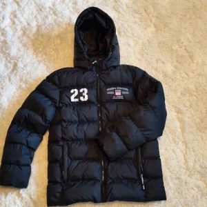 Svart pufferjacka från Vinson, strl 150 - Svart pufferjacka från Vinson med huva och dragkedja. Jackan har tryck med siffran 23 och Vinson Original-logga på bröstet. Quiltad design och två fickor med dragkedja. Perfekt för kalla dagar och riktigt snygg streetstil.