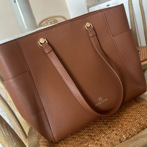 Brun shoppingväska från Anna Field - Stilren och rymlig shoppingväska från Anna Field i brun skinnimitation med guldfärgade detaljer. Väskan har två långa axelremmar och en enkel, elegant design som passar perfekt för dig som gillar klassisk stil. Perfekt storlek för att få plats med allt du behöver. I nytt skick då den aldrig är använd!🥰👏🏻