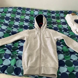 Ljusgrå hoodie från McKenzie XS - Snygg ljusgrå hoodie från McKenzie i storlek XS. Tröjan har dragkedja, huva med snörning och ribbade muddar. Perfekt för chill dagar eller när du vill ha en enkel och clean look. Materialet är mjukt och skönt, troligtvis bomullsmix.