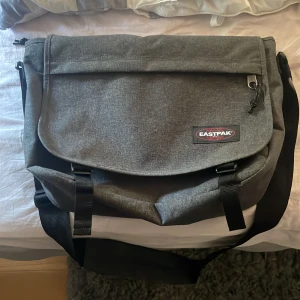 Grå datorväska från Eastpak - Snygg och praktisk grå datorväska från Eastpak med justerbar axelrem och klassisk logga på framsidan. Väskan har ett stort huvudfack, lock med spännen och dragkedja upptill. Perfekt för skola eller jobb när du vill ha med dig datorn.