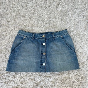 Blå jeanskjol med knappar fram - Snygg blå jeanskjol med kort längd och raka fickor fram. Kjolen har knappar längs hela framsidan, bälteshällor och dekorativa fickor bak. Midjemåttet är 37cm rakt över och kjolen är 29cm lång 💓