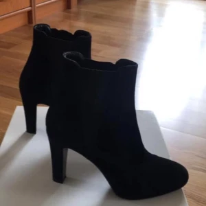   Rizzo Svarta mocka stövletter med klack - Snygga svarta stövletter i mocka med hög klack och rund tå. Skorna har en stilren och elegant design med diskret resår på sidorna för enkel påtagning. Perfekta för dig som vill ha en klassisk och trendig look.