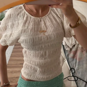 Vit ribbad blus från Gina Tricot - Supersöt vit blus från Gina Tricot i storlek XS. Toppen har ett ribbat, smockat tyg som ger en cool textur och sitter snyggt mot kroppen. Rund halsringning och korta puffärmar ger en fräsch vibe. Perfekt till jeans eller kjol för en chill look. Oanvänd endast påsatt för att visa hur den ser ut på💕