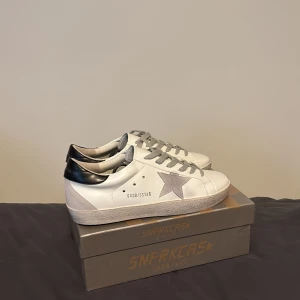 Golden Goose vita sneakers med stjärna - Golden Goose sneakers i vitt skinn med grå mockadetaljer och en stor grå stjärna på sidan. Skorna har grå snörning, svart hälparti och platt sula med mönstrad undersida. Snygg och clean design med GGDB/SSTAR-tryck på sidan. Perfekt för dig som gillar streetstyle.