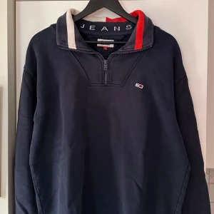 Marinblå half zip tröja Tommy Jeans - Snygg marinblå långärmad tröja från Tommy Jeans med half zip och cool krage med logotyp. Kragen har detaljer i rött, vitt och grått. Perfekt för dig som gillar streetwear och vill ha en chill look. Mjuk bomullskvalitet och relaxed passform.