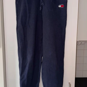 Mörkblå mjukisbyxor Tommy Jeans - Snygga mörkblå mjukisbyxor från Tommy Jeans med broderad logga på höften. Byxorna har snörning i midjan och är gjorda i mjukt bomullsmaterial. Perfekta för chill eller häng med kompisar. Klassisk loose passform och stilren design. Har ett litet hål framtill.