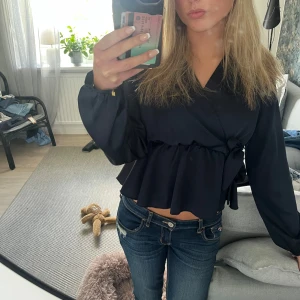 Svart omlottblus med volang - Säljer en svart omlottblus med långa ärmar och volang nertill. Blusen har en snygg v-ringning och knyt i sidan som ger en feminin touch. Perfekt till jeans för en trendig look.