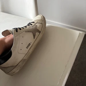Golden Goose Superstar sneakers vit/silver - Golden Goose Superstar sneakers i vitt skinn med silverfärgad stjärna på sidan och svart häl. Svarta snören och rund tå. Platt sula och cool vintage vibe. Perfekt för dig som gillar streetstyle och vill sticka ut med snygga detaljer. Dustbag, extra snören och box tillkommer! Tyvärr inte original sula.