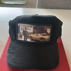 Svart Maggiore truckerkeps med motiv - Snygg svart truckerkeps från Maggiore med mesh på sidorna och bak, justerbar spänne och ett coolt motiv med bil och man på framsidan. Insidan har Maggiore-logga på banden. Perfekt för dig som gillar bilar och streetstyle.