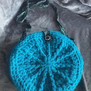 Turkos stickad axelväska med blommigt foder - Unik rund axelväska i turkos stickat material med blommigt tygfoder och matchande axelrem. Väskan stängs med en knapp. Mått 29x29 30kr+frakt