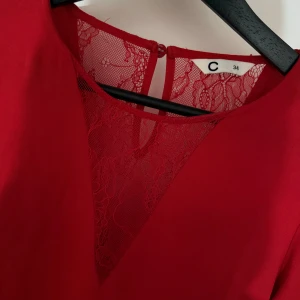 Röd blus med spetsdetalj från C - Röd långärmad blus med djup v-ringning och transparent spetsinfällning framtill. Blusen har en knapp i nacken och är tillverkad i ett lätt, flowy material som ger en snygg siluett. Perfekt för dig som vill sticka ut med en färgstark och elegant look.