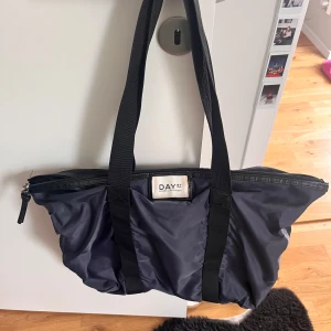 Svart axelväska från DAY  - Stor och rymlig svart axelväska från DAY ET Birger et Mikkelsen. Tillverkad i glansig syntet med långa svarta band och dragkedja upptill. Perfekt för att få plats med allt du behöver, både till skolan och på språng.