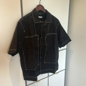 Svart overshirt från Zara med kontrastsömmar - Helt oanvänd! Svart overshirt från Zara med vita kontrastsömmar och stora fickor framtill. Skjortan har korta ärmar, klassisk krage och knäppning med svarta knappar. Perfekt för en stilren och edgy look. Materialet känns som bomull och passformen är normal.