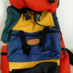 Färgglad ryggsäck från Twinings - Stor och färgglad ryggsäck från Twinings med flera fack och dragkedjor. Väskan har paneler i rött, blått, gult, grönt och svart och är tillverkad i slitstark polyester. Perfekt för dig som vill sticka ut och ha plats för allt du behöver på äventyret.