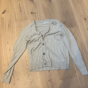 Beige kofta från Jack and Jones med knappar - Beige kofta från Jack and Jones med långa ärmar och stora knappar framtill. Enkel och stilren design i mjukt bomullsmaterial, perfekt att slänga över en t-shirt. Ribbstickade muddar vid ärmslut och nederkant för en snygg passform.