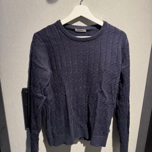 Mörkblå stickad tröja J.Lindeberg - Snygg mörkblå stickad tröja från J.Lindeberg med coolt geometriskt mönster. Tröjan har rund halsringning, ribbade muddar och långa ärmar. 
