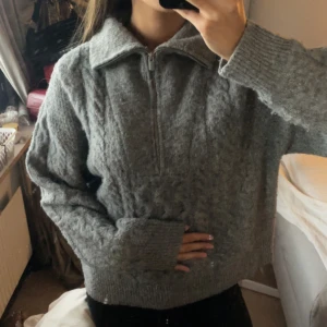 Grå stickad half zip tröja - Mysig grå stickad tröja med half zip och stor krage. Tröjan har ett snyggt kabelstickat mönster framtill och långa ärmar med breda muddar. Perfekt för lager-på-lager och chill dagar.