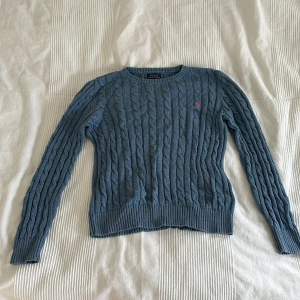 Blå kabelstickad tröja Polo Ralph Lauren - Kabelstickad blå tröja från Polo Ralph Lauren med rund halsringning och lång ärm. Klassisk design med rosa broderad logga på bröstet. Perfekt till jeans eller kjol.