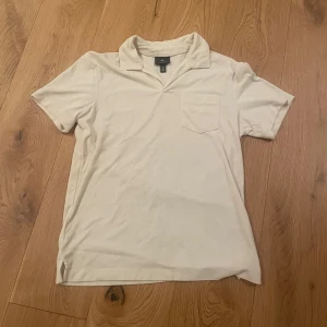 Vit frótte piké från H&M - Vit piké t-shirt från H&M med klassisk krage och bröstficka. Regular fit och kortärmad modell i mjuk bomull, perfekt för en clean och enkel stil. Passar dig som gillar basic men ändå vill ha något med lite extra detaljer.