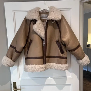 Beige teddyjacka från Stand Studio - Säljer en beige teddyjacka från Stand Studio med mjukt fuskpälsfoder, bred krage och detaljer i mörkbrunt. Jackan har dragkedja framtill, stora fickor och breda muddar i teddy. Perfekt för hösten eller kalla dagar och riktigt snygg till jeans eller kjol.