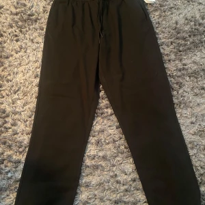 Svarta byxor  från Les Deux - Säljer ett par svarta mjukisbyxor från Les Deux, modell Patrick Mélange Pants. Byxorna har en relaxed passform med snörning i midjan och är gjorda i mjukt material som är perfekt för chill dagar. Snyggt stilrena och passar till sneakers. Prislappen finns fortfarande kvar 