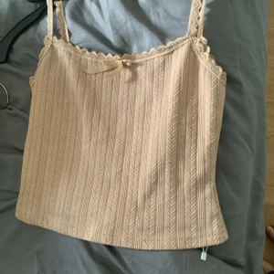  Topp  - Supersöt beige linnetopp med smala axelband och spetskant runt om. Toppen har ett diskret ribbat mönster med vertikala detaljer och en liten rosett framtill. Perfekt att styla med jeans eller kjol för en chill look.