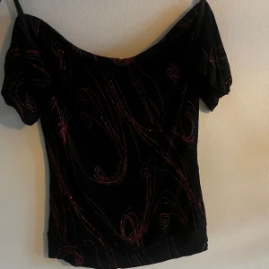 Svart offshoulder topp  - Snygg svart offshoulder topp med smala axelband. Toppen har ett glittrigt rött mönster i virvlande linjer som ger en cool och unik look. Perfekt för fest eller middag med vänner! Tröjan har öppen rygg!