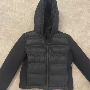 Svart pufferjacka från Zara  - Svart pufferjacka från Zara med huva och dragkedja framtill. Jackan har quiltad front och rygg i glansigt material, medan ärmarna är släta och matta. Använd bara några gånger. Perfekt för kyliga dagar och riktigt snygg till streetwear-stilen.