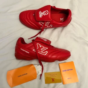 Louis Vuitton röda fotboll sneakers - Unika röda fotbollsskor från Louis Vuitton med stora vita LV-loggor på sidan och tungan. Snygg design med snörning och platt sula, tillverkade i skinn. Tungan har coolt 'LV Players United'-tryck och yttersulan har mönstrade detaljer. Kommer med originalförpackning och extra skosnören.