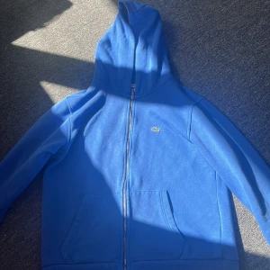 Blå hoodie från Lacoste med dragkedja - Blå hoodie från Lacoste med klassisk krokodillogga på bröstet. Tröjan har huva, hel dragkedja och två stora fickor framtill. Mjuk insida och ribbade muddar vid ärmslut och nederkant. Perfekt för chill dagar eller när du vill ha en clean look.