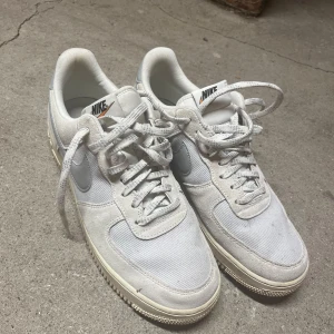 Nike Air Force 1 sneakers - Nike Air Force 1 sneakers i vit/beige/grå färg med klassisk rund tå och platt sula. Skorna har snörning och är tillverkade i en mix av skinn och syntetmaterial. Snygg retrostil med ikonisk Nike-logga på sidan och tungan. Perfekt för dig som gillar clean och stilren look.