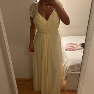 Ljusgul långklänning med volang - Superfin ljusgul långklänning med omlott framtill, v-ringning och volangdetaljer på axlarna. Klänningen har öppen rygg med knytband och faller snyggt längs kroppen. Perfekt för sommarens speciella tillfällen! Bra skick, använt en gång på ett bröllop, säljer för att den är för stor samt att jag inte kommer använda den mer.