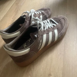 Adidas Originals bruna Gazelle sneakers - Bruna Adidas Gazelle sneakers i mocka med klassiska vita ränder och vit häl. Skorna har vit snörning och en gummisula i ljusbrun ton. Perfekta för dig som gillar retrovibbar och streetstyle. Ikonisk Adidas-logga på sidan och innersulan.