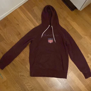 Vinröd hoodie från Gant - Säljer en snygg vinröd hoodie från Gant med ett tryckt emblem på bröstet. Hoodien har en klassisk design med känguruficka och justerbar huva med snören. Tillverkad i en bomullsblandning för extra komfort.