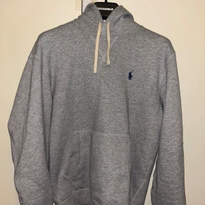 Grå hoodie från Ralph Lauren - Säljer en klassisk grå hoodie från Ralph Lauren med en liten blå logga på bröstet. Tröjan har en justerbar huva med vita snören och en stor magficka framtill. Perfekt för en avslappnad stil.10/10 skick, knappt använd! 